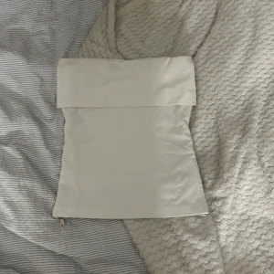 Vit axelbandslös tubtopp med dragkedja - Säljer en stilren vit tubtopp med bred kant upptill och en snygg silverfärgad dragkedja längs sidan. Toppen är axelbandslös och har en clean, minimalistisk look som passar perfekt till jeans eller kjol. Materialet känns slätt och har en lätt glans.