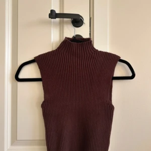Brun ribbad ärmlös polotopp - Säljer en brun ribbad polotopp med hög hals och ärmlös design. Toppen har en tight passform och är stickad i ett mjukt material som ger en snygg siluett. Perfekt att styla med jeans eller kjol för en trendig look. Inga defekter och knappt använd 