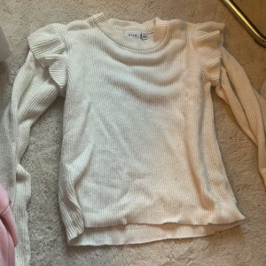 Vit ribbad stickad tröja från Vila - Supersöt vit stickad tröja från Vila med ribbad struktur och volangdetaljer på axlarna. Långärmad och med rund halsringning, perfekt för lager-på-lager under hösten och vintern. Enkel att styla till både jeans och kjol.