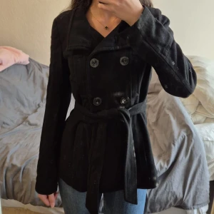 Svart kappa med bälte och knappar - Stilren svart kappa med dubbelknäppning och bälte i midjan. Jackan har långa ärmar, klassisk krage och stora knappar framtill. Perfekt för dig som gillar en tidlös look med lite edge. Materialet känns mjukt och värmande, passar bra till jeans eller klänning. Passar mig som har xs/s i jackor också. 