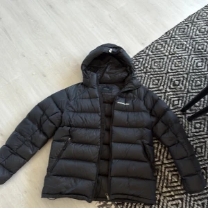 Svart pufferjacka från Peak Performance - Säljer en svart pufferjacka från Peak Performance med huva och dragkedja. Jackan har en quiltad design, två sidofickor med dragkedja och logga på bröstet. Perfekt för kalla dagar och riktigt skön att bära. Har fått ett litet hål på ryggen som är ihopsytt. Använde under hela förra vintern men har köpt nu vinterjacka nu därför den är på försäljning. De ihopsydda på ryggen syns ej. Otroligt skön och varm.