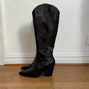 Mörkbruna cowboy boots från ALDO - Aldrig använda. Snygga svarta cowboy boots från ALDO i äkta skinn med präglad struktur som ger en cool reptilkänsla. Boots med spetsig tå, hög klack och klassisk western-inspirerad form. Perfekta för dig som vill sticka ut med en edgy stil. Finns ett par i färgen ”other black” i samma kollektion (Nevada) ute på Zalando för 1 995 kr.