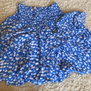 Blå blommig volangkjol i storlek S - Supersöt blå kortkjol med vita blommor och volanger. Kjolen har smock i midjan för en bekväm passform och är perfekt för varma dagar. Lätt och luftig, passar dig som gillar en feminin och somrig stil.