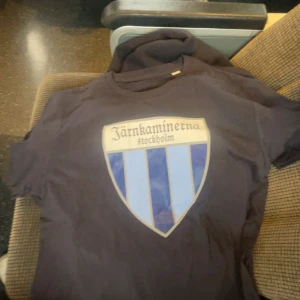 Järnkaminerna t-shirt blå S - Mörkblå t-shirt med stort Järnkaminerna Stockholm-tryck på bröstet. Klassisk rund hals och korta ärmar. Perfekt för dig som vill visa ditt stöd för laget. Skön och mjuk bomullskänsla. Mått 62 x 43