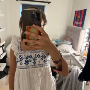 Vit och blå broderad sommarklänning - Superfin vit klänning med blå broderier och volangdetaljer på axlarna. Klänningen har knytning i ryggen och är ärmlös med en luftig passform. Perfekt för varma dagar och har en söt, romantisk vibe.