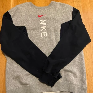 Nike grå sweatshirt med svarta ärmar - Nike sweatshirt i grått med svarta ärmar och röd logga på bröstet. Tröjan har klassisk passform, rund hals och är tillverkad i mjuk bomullsblandning. Perfekt för en sportig och avslappnad stil.