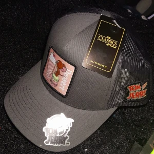 Tom & Jerry Keps  - Snygg grå truckerkeps med Tom & Jerry-motiv och dessertpatch framtill. Kepsen har svart mesh baktill för extra ventilation och justerbar snapback. 