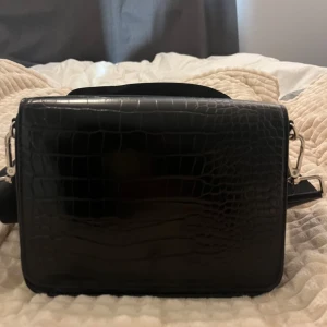 Svart axelväska i croco-mönster - Snygg svart axelväska med croco-mönstrad yta och bred justerbar axelrem. Väskan har silverfärgade metalldetaljer och en stilren, boxig form som ger en modern vibe. Perfekt för dig som vill ha en trendig och praktisk väska till vardagen. Använd men inget som märks🩵