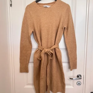 Beige stickad klänning med bälte - Mysig beige stickad klänning med lång ärm och rund hals. Klänningen har ett matchande knytbälte i midjan.