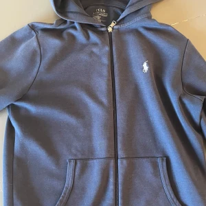 Mörkblå hoodie från Polo Ralph Lauren - Hej! Säljer en snygg hoodie från Polo Ralph Lauren med dragkedja och huva.  Tröjan har klassisk logga broderad på bröstet, snörning i huvan och praktiska fickor framtill.  Tillverkad i mjuk bomull och polyester för skön komfort.  Perfekt för chill dagar eller när du vill ha en clean look. Den är pyttelite solblekt nertill, men man ser knappt det.   Vill du veta något mer eller vill ha fler bilder så är det bara att skriva! Priset är såklart inte hugget i sten!