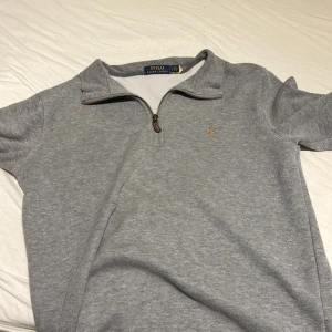 Grå half zip tröja från Polo Ralph Lauren - Säljer en stilren grå tröja från Polo Ralph Lauren med klassisk broderad logga på bröstet. Tröjan har half zip-dragkedja och är tillverkad i mjuk bomull. Perfekt för lager-på-lager och chill vardagsstil.