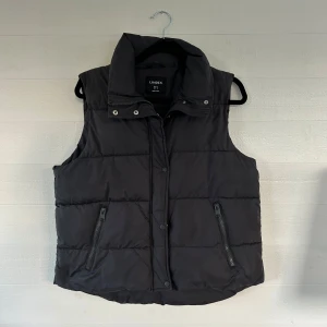 Svart dunväst - Svart quiltad dunväst från Lindex med hög krage och två dragkedjefickor framtill. Västen har en stilren look med raka linjer och stängs med både dragkedja och tryckknappar. Perfekt för lager-på-lager under kyliga dagar.🤩💕
