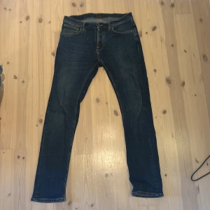 Mörkblå jeans från Nudie Jeans Co W33 L34 - Snygga mörkblå jeans från Nudie Jeans Co med klassiska orange sömmar och broderade bakfickor. Modellen har rak passform och normal midja. Tillverkade i slitstarkt denim med detaljer som läderpatch bak i midjan. Bara en defekt som sitter vid höger fram ficka men inget som märks av vid användning.