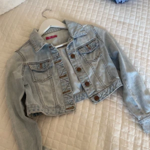 Ljusblå croppad jeansjacka från YD - Snygg ljusblå croppad jeansjacka från YD med klassisk krage, bruna knappar och två bröstfickor med lock. Jackan har långa ärmar och coola sömdetaljer. Perfekt för dig som vill ha en chill och trendig look.