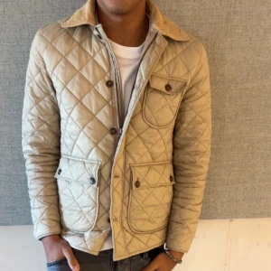 Beige quiltad jacka These Glory Days - Snygg beige quiltad jacka från These Glory Days med klassisk krage i manchester, tre fickor framtill och två innefickor. med knappar och stilrena detaljer. Perfekt till hösten men funkar också bra på vintern. Modellen är 180