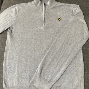 Grå half zip tröja från Lyle & Scott - Snygg grå långärmad tröja från Lyle & Scott med klassisk gul logga på bröstet. Tröjan har en half zip-dragkedja och ribbade muddar vid ärmslut och nederkant. Perfekt för dig som gillar stilren och enkel design.
