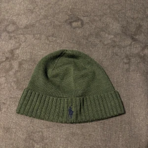 Olivgrön mössa från Polo Ralph Lauren - Snygg olivgrön mössa från Polo Ralph Lauren i mjuk och tvättbar merinoull. Klassisk ribbstickad design med uppvikt kant och broderad logga framtill. Perfekt accessoar för kyliga dagar och enkel att matcha med din höst- eller vinterstil.