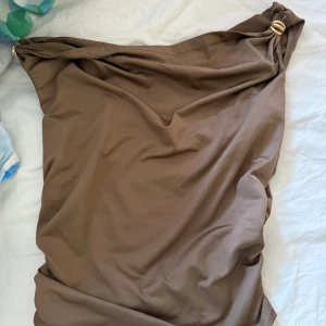 Två axelbandslösa klänningar med gulddetalj - Säljer två stilrena axelbandslösa toppar, en i brun och en i vit. Båda har snygga guldfärgade ringdetaljer vid axeln och är sydda i ett mjukt, stretchigt material. Perfekt för dig som gillar minimalistisk och trendig look.