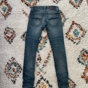 Nudie Jeans tighta blå jeans 24/30 - Säljer ett par tighta blå jeans från Nudie Jeans i storlek 24/30. Jeansen har klassisk femficksdesign, slitningar på låren och knän, samt Nudie-logga på bakfickorna. De är i stretchigt denim och har en snygg tvättad look.