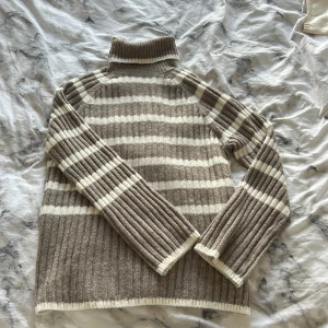 Randig grå/beige stickad polotröja - Supermysig polotröja med grå/beige och vita ränder. Tröjan är ribbstickad och har lång ärm samt hög krage som kan vikas ner. Slits på båda sidorna. (Helt oanvänd) 