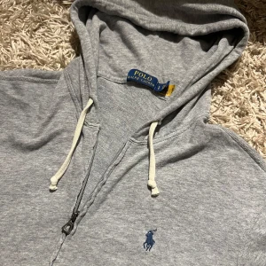 Grå hoodie från Polo Ralph Lauren - Snygg grå hoodie från Polo Ralph Lauren med klassisk blå broderad logga på bröstet. Tröjan har huva med vita snören, dragkedja framtill och två fickor. Postas antigen samma dag som köp eller dagen efter. Mvh priset är inte hugget i sten 