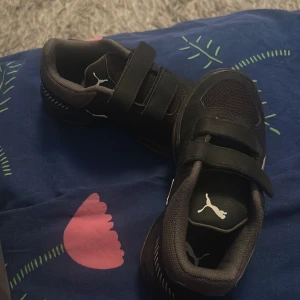 Svarta sneakers från Puma med kardborre - Säljer ett par svarta sneakers från Puma med vita detaljer och bruna sulor. Skorna har två breda kardborreband för enkel påtagning och meshpartier för extra andningsförmåga. Perfekta för dig som vill ha både stil och funktion i vardagen.