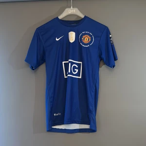 Manchester United Ronaldo 7 blå tröja - Blå Manchester United fotbollströja från Nike med Ronaldo 7 på ryggen. Tröjan har korta ärmar, vitt tryck och klubbmärke på bröstet. Materialet är lätt och andas, perfekt för match eller träning. Snygg detalj med IG-logga och FIFA-märke framtill.