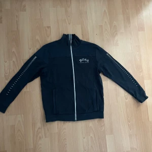 Svart trackjacka från Hugo Boss - Svart trackjacka från Hugo Boss med vit dragkedja och logga på bröstet. Jackan har ståkrage, långa ärmar med vita streckdetaljer längs sidorna och två fickor framtill. Tillverkad i mjuk bomullsmix för en skön och sportig look.