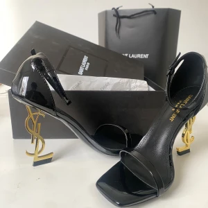 Saint Laurent svarta sandaler med YSL-klack - Exklusiva svarta sandaler från Saint Laurent i lackat skinn med öppen tå och smal rem över foten. Klacken är i guld och formad som YSL-loggan, vilket ger en riktigt lyxig vibe. Snygg fyrkantig tå och justerbar rem runt ankeln. Perfekta för dig som vill sticka ut.