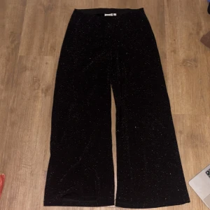 Svarta glittriga byxor från Kappahl - Svarta byxor från Kappahl med glittrigt tyg och bred resår i midjan. Byxorna har en rak och vid passform 