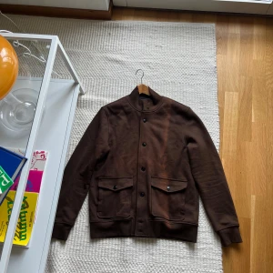 Brun bomberjacka/tröja från Morris - Snygg brun bomberjacka/tröja från Morris med ribbad krage och muddar. Jackan har två stora fickor framtill med lock och knappar, samt stängs med knappar hela vägen. Perfekt för dig som vill ha en stilren och klassisk look. Jackan/tröjan är använd max 5 gånger men har fått blivit solblekt (se bild) 