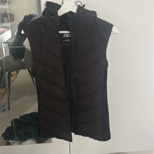 Svart väst Zara - Säljer en svart dunväst från Zara med huva och dragkedja framtill. Västen har en quiltad design med sneda sömmar och är lite längre i modellen. Perfekt att slänga över en hoodie eller tröja för en cool och avslappnad look.