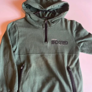 Grön hoodie från Blue Seven med huva - Mysig grön hoodie från Blue Seven med trycket 'Sound Vibes' på bröstet. Tröjan har huva, halv dragkedja framtill och två fickor med dragkedja. Perfekt för kyliga dagar och enkel att matcha med jeans eller joggers.