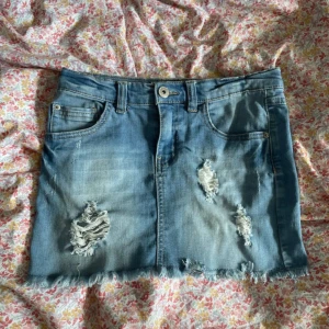 Blå jeanskjol från LAB Industries - Snygg ljusblå jeanskjol från LAB Industries med slitna detaljer och fransad nederkant. Klassisk femficksmodell med knapp och dragkedja framtill. Perfekt för dig som gillar en avslappnad och trendig stil.