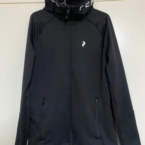 Svart jacka från Peak Performance - Säljer en stilren svart jacka från Peak Performance. Den har en dragkedja framtill och en hög krage med texten 'PEAK PERFORMANCE'. Jackan är långärmad och perfekt för både träning och vardag. Materialet känns slitstarkt och bekvämt.