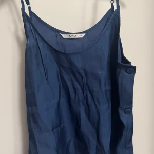Mörkblå linne från ONLY - Säljer en stilren mörkblå linne från ONLY med justerbara smala axelband och dekorativa knappar på sidan. Toppen har en lätt glansig finish och är perfekt för att skapa en enkel men snygg look. Passar till både jeans och kjol.