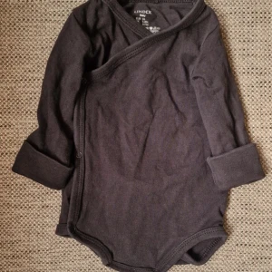 Svart omlottbody från Lindex Baby - Mysig svart omlottbody från Lindex i mjuk bomull. Långärmad modell med praktiska tryckknappar längs sidan och vid grenen. Perfekt för de allra minsta och enkel att ta på och av. 95% bomull, 5% elastan.