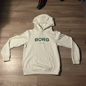 Vit hoodie från Björn Borg - Vit hoodie från Björn Borg med grön BORG-logga på bröstet. Klassisk huva med snörningsdetaljer och stor magficka framtill. Mjuk och skön, perfekt för chill dagar. Långärmad och i bomullsmix för extra komfort.