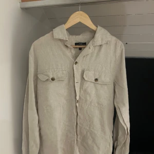 Beige linneskjorta från Hampton republic - Snygg beige skjorta i linne från Tailored Originals. Skjortan har två bröstfickor med knappar, klassisk krage och långa ärmar. Perfekt för en avslappnad men stilren look under varmare dagar.