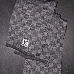 Louis Vuitton halsduk - Snygg halsduk från Louis Vuitton i klassiskt rutigt mönster. Färgen är en mix av svart och grått, vilket gör den lätt att matcha med olika outfits. Halsduken har en vävd LV-logga och känns både lyxig och stilren.