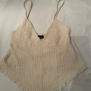 Krämvit linnetopp från Gina Tricot - Supersöt krämvit linnetopp från Gina Tricot med smala flätade axelband och v-ringning. Toppen har en strukturerad, rynkad yta och är lätt utsvängd nertill. Perfekt för en fräsch och somrig look.
