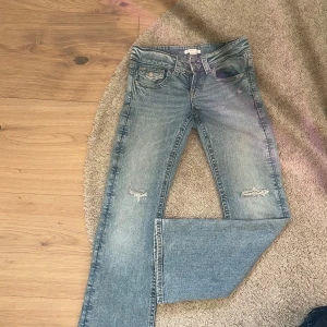 Blå slitna jeans från Gina Tricot - Ett par ljusblå jeans från Gina Tricot med snygga slitningar vid knäna och klassisk femficksdesign. Jeansen har normal passform och är tillverkade i bomullsmaterial. Perfekta för en avslappnad och trendig look.