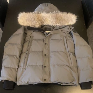 Grå dunjacka med fuskpäls från Canada goose - Snygg grå dunjacka från Canada goose med avtagbar huva och fuskpäls. Jackan har quiltad design, dragkedja och knappar framtill samt två stora fickor. Perfekt för kalla dagar och riktigt varm.