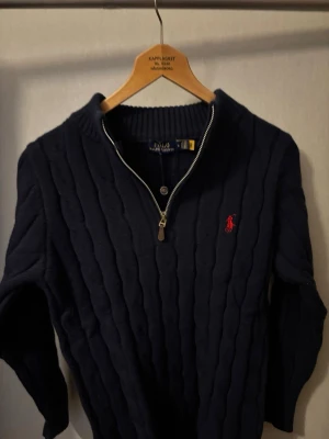 Mörkblå stickad tröja Polo Ralph Lauren - Mörkblå stickad tröja från Polo Ralph Lauren med halv dragkedja och ribbad krage. Klassisk kabelstickad design och röd broderad logga på bröstet. Perfekt för lager på lager och en stilren look.