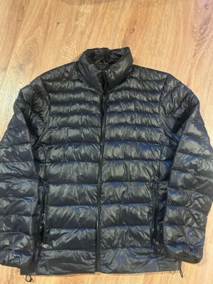 Polo Ralph Lauren Perfromance Jacka - Snygg svart dunjacka från Polo Ralph Lauren med quiltad design och hög krage. Jackan har dragkedja framtill och två sidofickor med dragkedja. Perfekt för kyliga dagar och enkel att matcha med olika outfits. 