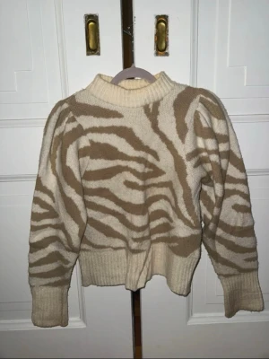 Beige zebramönstrad stickad tröja - Säljer en beige och vit stickad tröja med zebramönster. Tröjan har puffärmar, bred ribbad mudd vid ärmslut och midja samt hög hals. Perfekt för dig som vill ha en trendig och mysig look med djurmönster.