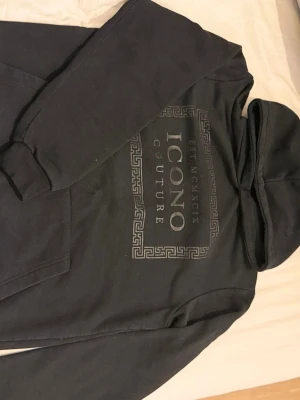 Svart hoodie från Icono Couture - Svart hoodie från Icono Couture med stor logga och dekorativt mönster på bröstet. Klassisk huva och magficka framtill. Tillverkad i mjukt material som känns skönt mot huden. Perfekt för en avslappnad och trendig look.