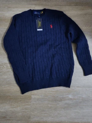 Mörkblå kabelstickad tröja Polo Ralph Lauren - Kabelstickad mörkblå tröja från Polo Ralph Lauren med klassisk röd logga broderad på bröstet. Tröjan har rund halsringning, ribbstickade muddar och är tillverkad i mjukt material. Perfekt för dig som gillar stilrena och tidlösa plagg.