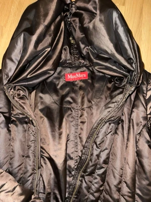 Brun lång dunjacka från Max Mara - Snygg lång brun dunjacka från Max Mara med quiltad design och stor huva. Jackan har dragkedja framtill och en glansig finish som ger ett exklusivt intryck. Perfekt för kalla dagar när du vill hålla dig varm och se stilren ut.