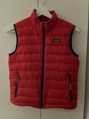 Röd dunväst från Patagonia - Snygg röd dunväst från Patagonia med quiltad design och marinblå detaljer vid dragkedja och ärmöppningar. Västen har två framfickor med dragkedja och står upp i kragen för extra värme. Perfekt lager på lager-plagg för kyliga dagar.